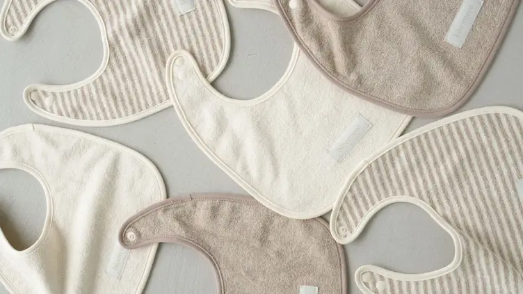 Vários babadores de tecido em tons neutros de bege e branco com estampas listradas e lisas dispostos sobre fundo cinza