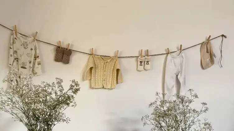 Roupas de bebê penduradas em um varal de corda contra uma parede branca com ramos de flores nas laterais