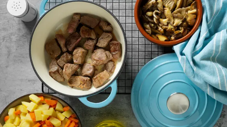 Panela de ferro azul com pedaços de carne, ao lado de tigelas com cogumelos, batatas e cenouras picadas