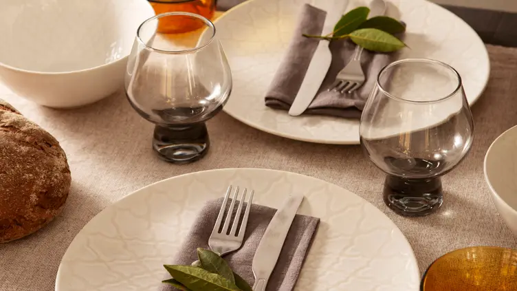 Mesa posta com pratos brancos, talheres, taças de vidro cinza e guardanapos de tecido sobre toalha de mesa bege