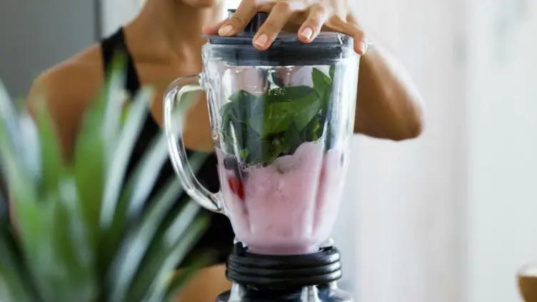 Pessoa segurando a tampa de um liquidificador com vitamina rosa e folhas verdes