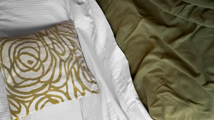 Cama com lençol branco, almofada branca com estampa geométrica dourada e cobertor verde musgo dobrado