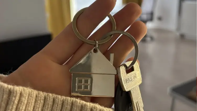 Mão segurando um chaveiro de metal em formato de casa junto com chaves