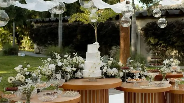 Mesa de casamento ao ar livre com bolo branco de quatro andares, flores brancas, doces e globos de vidro suspensos