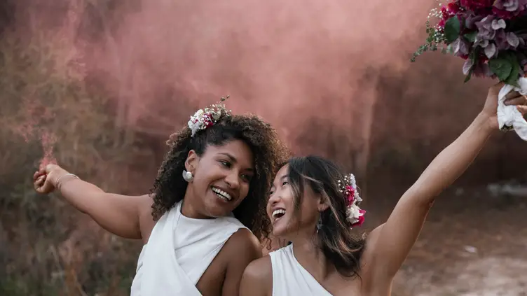 Duas noivas sorrindo com flores no cabelo, segurando um buquê e uma bomba de fumaça rosa ao ar livre