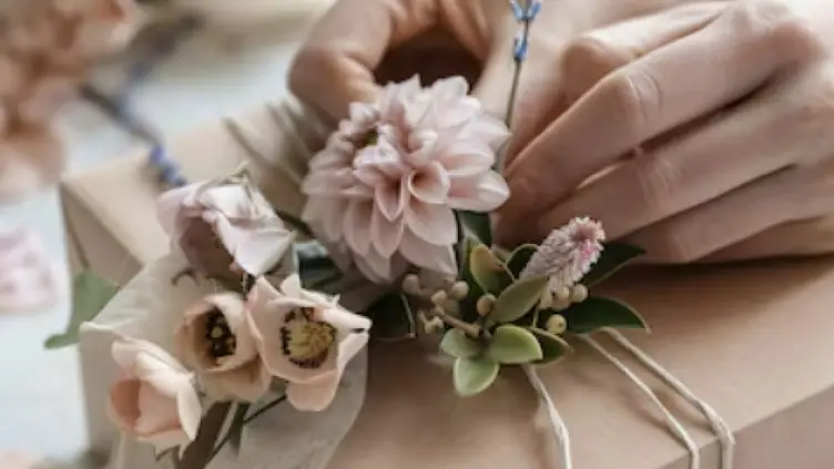 Mãos ajustando um arranjo de flores delicadas preso com barbante em uma caixa de presente embrulhada em papel pardo