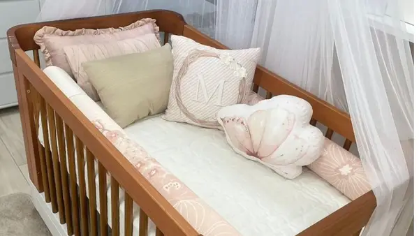 Berço de madeira com enxoval em tons de rosa e bege, contendo almofadas decorativas e um mosquiteiro ao fundo