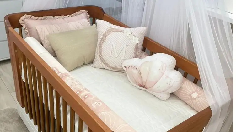 Berço de madeira com enxoval em tons de rosa e bege, contendo almofadas decorativas e um mosquiteiro ao fundo