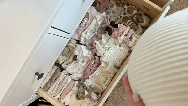Gaveta de cômoda branca aberta com roupas de bebê, sapatinhos, laços e um chocalho de crochê com barriga de grávida ao lado