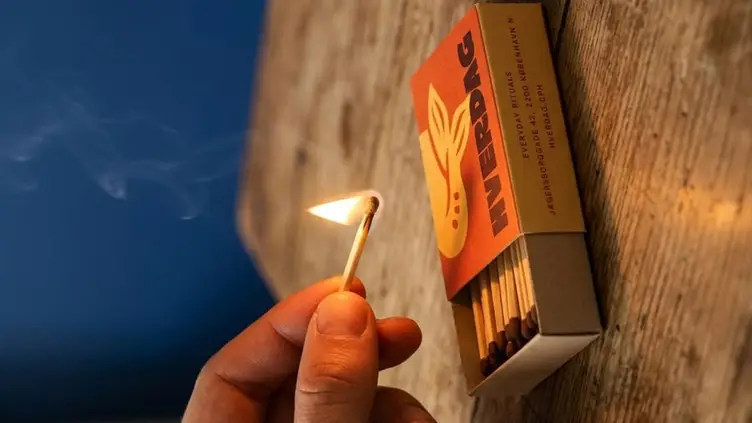 Mão segurando um palito de fósforo aceso ao lado de uma caixa aberta com a marca Hverdag sobre uma mesa de madeira